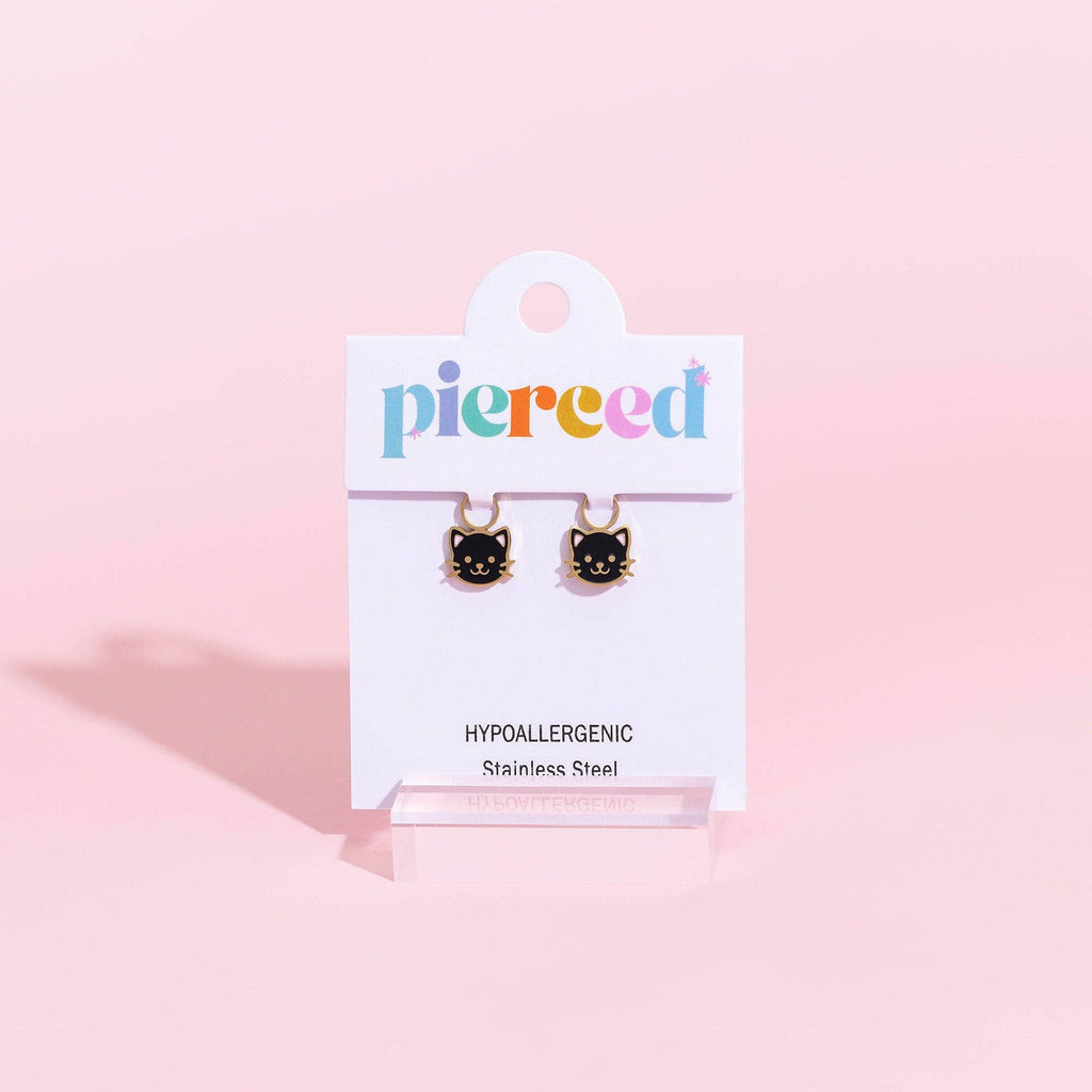 A pair of small black cat face stud earrings with gold-tone tops, displayed on a white card labeled 'pierced' against a soft pink background.