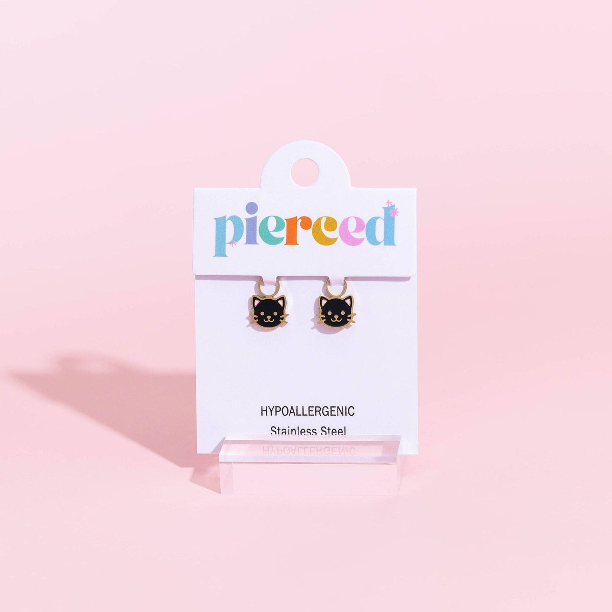 A pair of small black cat face stud earrings with gold-tone tops, displayed on a white card labeled 'pierced' against a soft pink background.