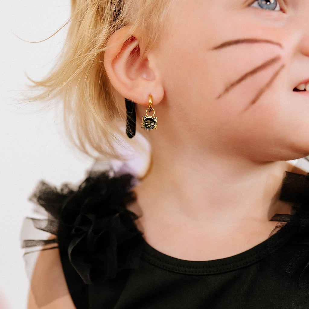 A close-up of a child's ear wearing a small gold hoop earring with a black cat-shaped charm featuring a green stone, against a plain white background.