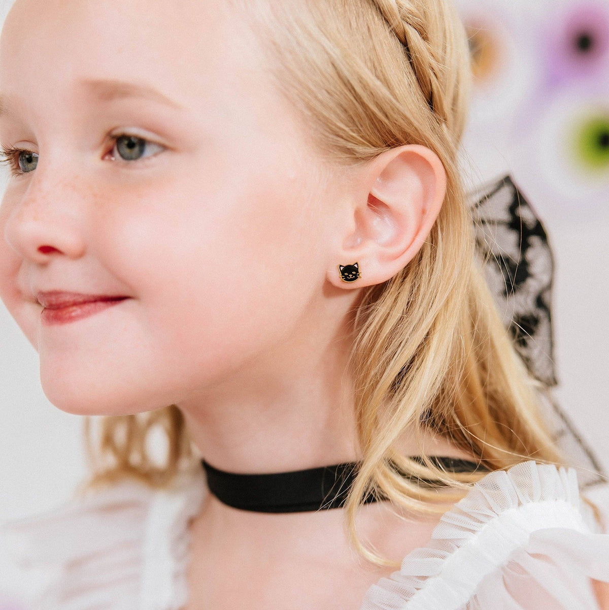 A close-up lifestyle shot of a young blonde girl wearing a black choker necklace and a small black stud earring with a clear center stone.