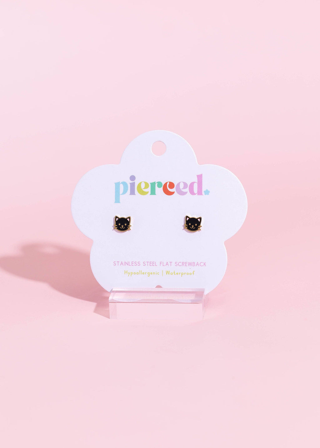 A pair of black cat-shaped stud earrings displayed on a white cloud-shaped card that reads 'pierced.' in colorful lowercase letters, set against a solid pastel pink background.