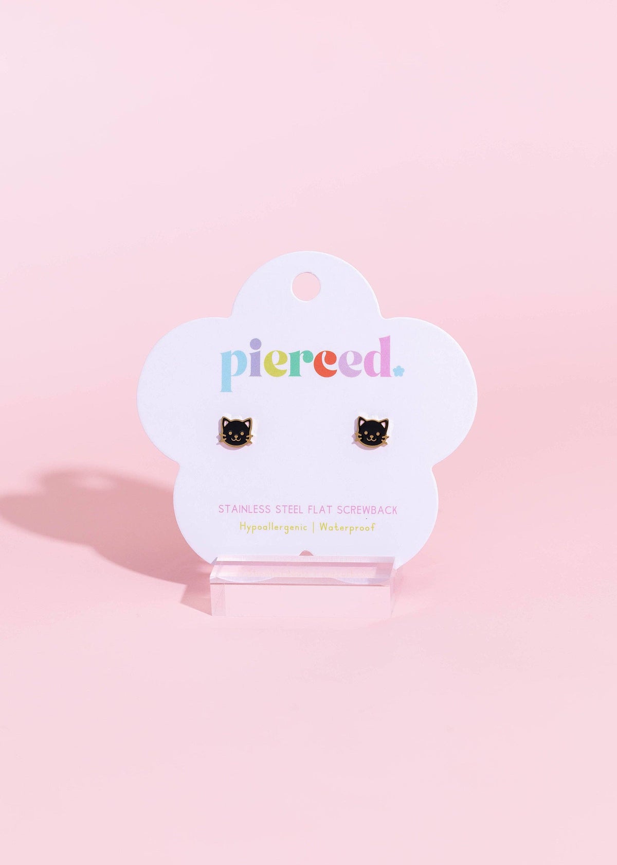A pair of black cat-shaped stud earrings displayed on a white cloud-shaped card that reads 'pierced.' in colorful lowercase letters, set against a solid pastel pink background.
