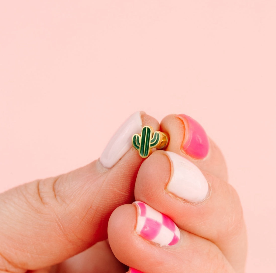 Hand holding a small green cactus-shaped earring against a pink background