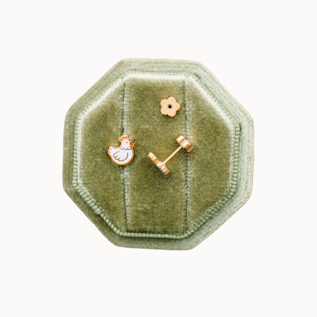 A pair of gold-toned stud earrings displayed on an octagonal sage green velvet jewelry box against a plain white background. One earring features a small white enamel rooster, and the other is a small gold flower with a post.
