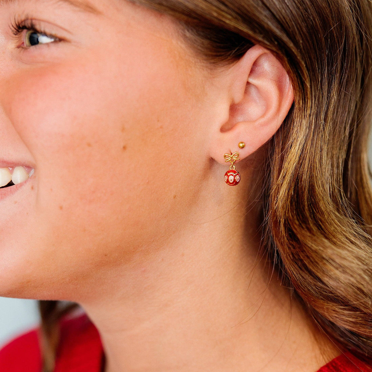 A close-up side profile of a woman wearing a small gold dangle earring with a red and white patterned bead, paired with a red knit sweater.