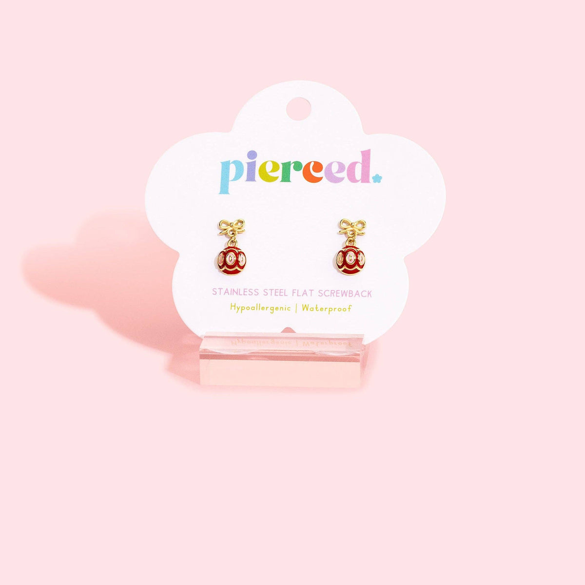 A pair of gold-tone flat screwback earrings with red and white enamel details, displayed on a white cloud-shaped pierced co brand card stand against a solid pink background.