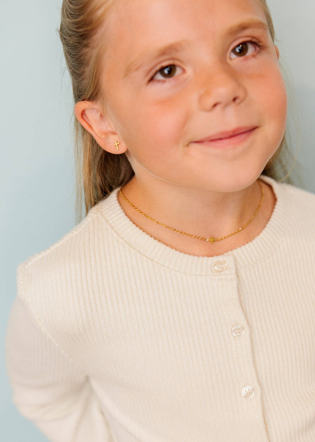 A young girl with blonde hair tied back wears a delicate gold chain necklace and matching small gold stud earrings, paired with a cream ribbed cardigan against a light blue background.