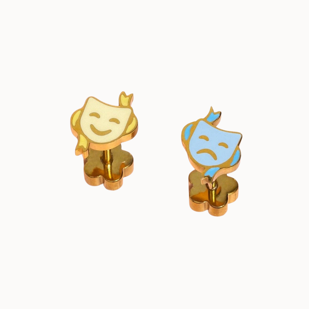 A pair of small enamel pin earrings shaped like comedy and tragedy masks on gold-tone backing, shown on a plain light background.