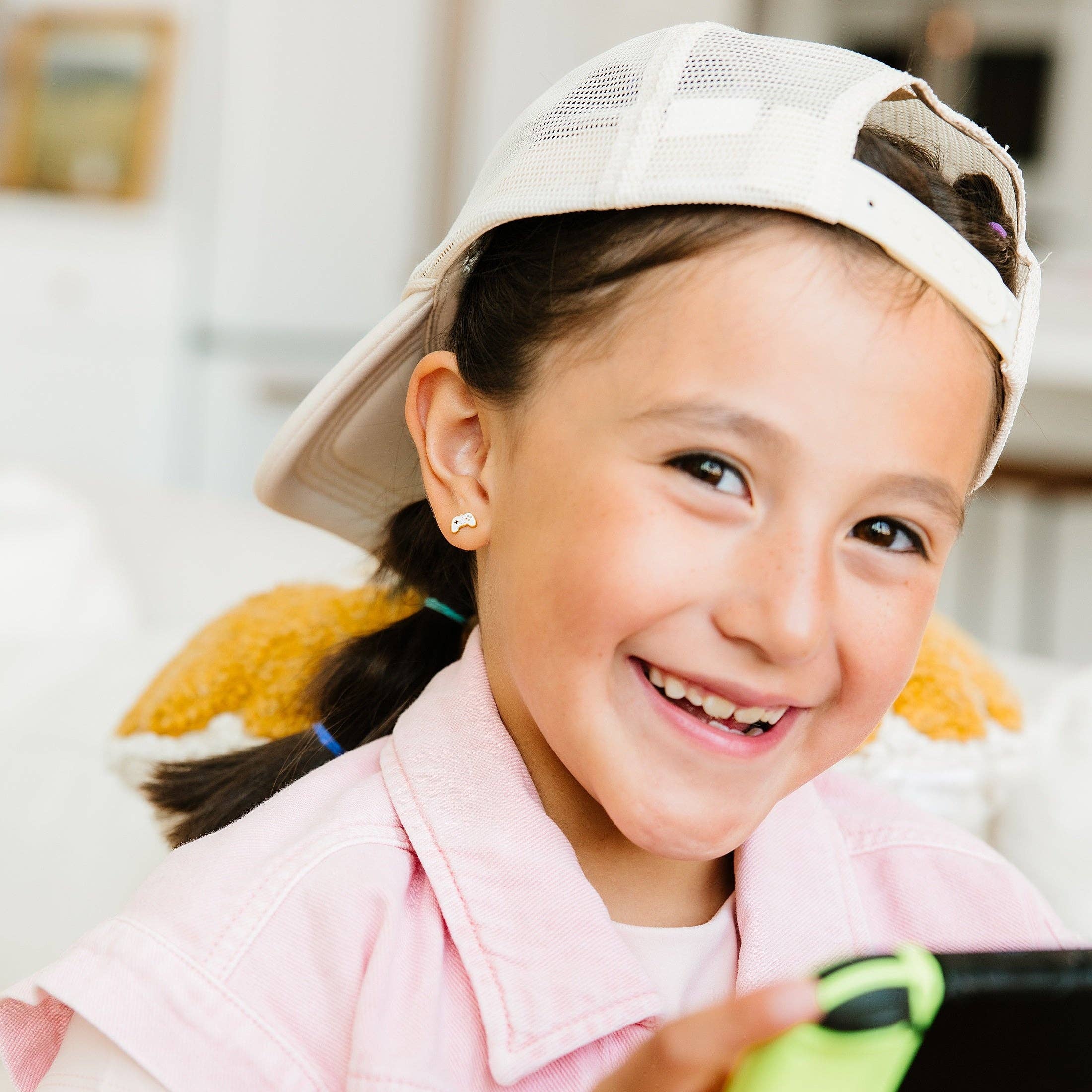 A young girl smiling while wearing a light pink denim-style shirt and a white baseball cap backwards, holding a black smartphone with a lime green case in a brightly lit indoor setting.