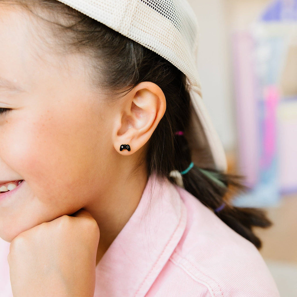 A close-up lifestyle shot of a young girl wearing a small black heart-shaped stud earring, a pink shirt, and a cream-colored mesh-back baseball cap.