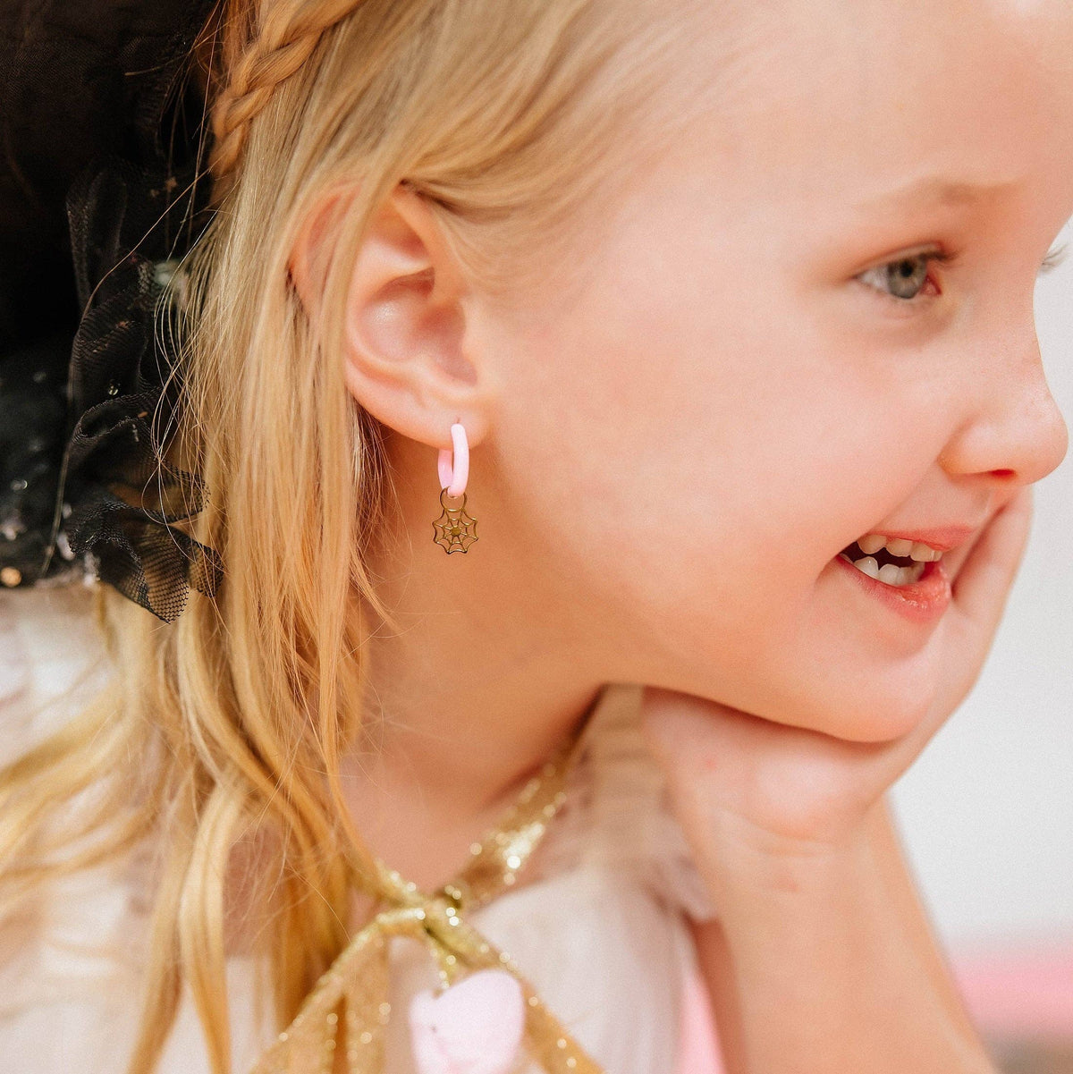 A close-up lifestyle shot of a young blonde girl wearing a pink hoop earring with a dangling gold fairy charm, looking off-camera with a smile.