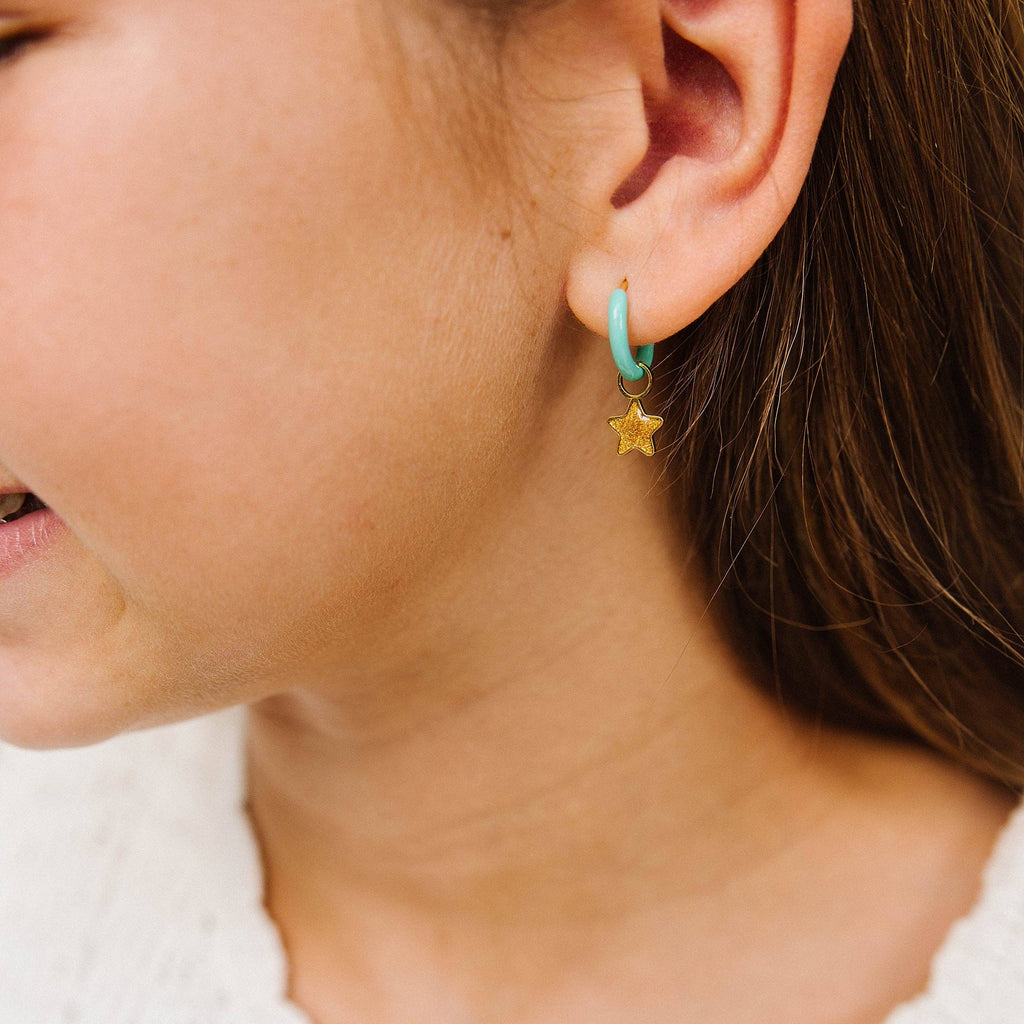A close-up of a person's ear wearing a small turquoise hoop earring with a dangling gold star charm, paired with a white knit top.