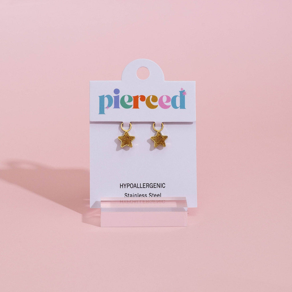 Gold star-shaped stud earrings on a white display card with the brand name 'pierced' in multicolored letters, set against a solid pink background.