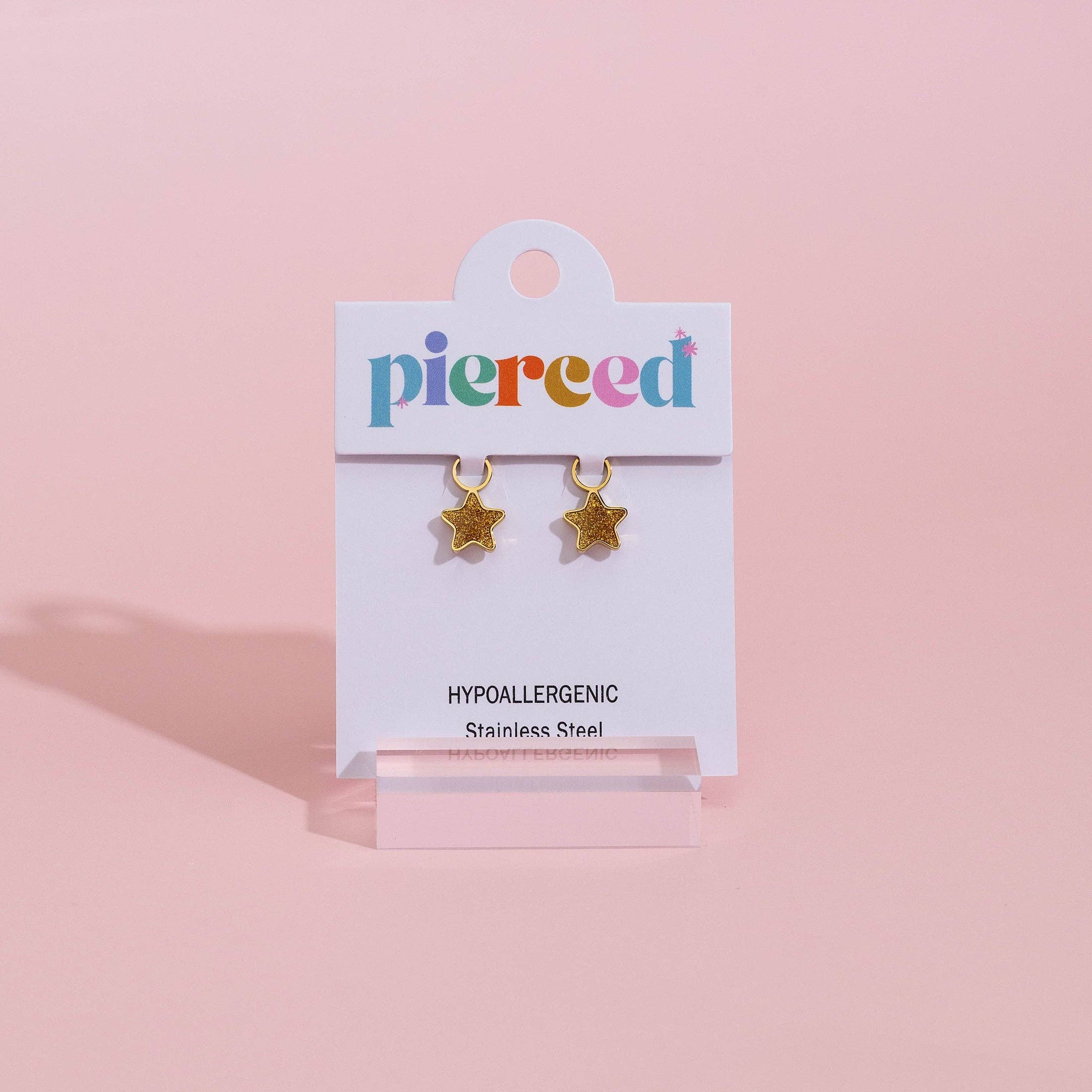 Gold star-shaped stud earrings on a white display card with the brand name 'pierced' in multicolored letters, set against a solid pink background.
