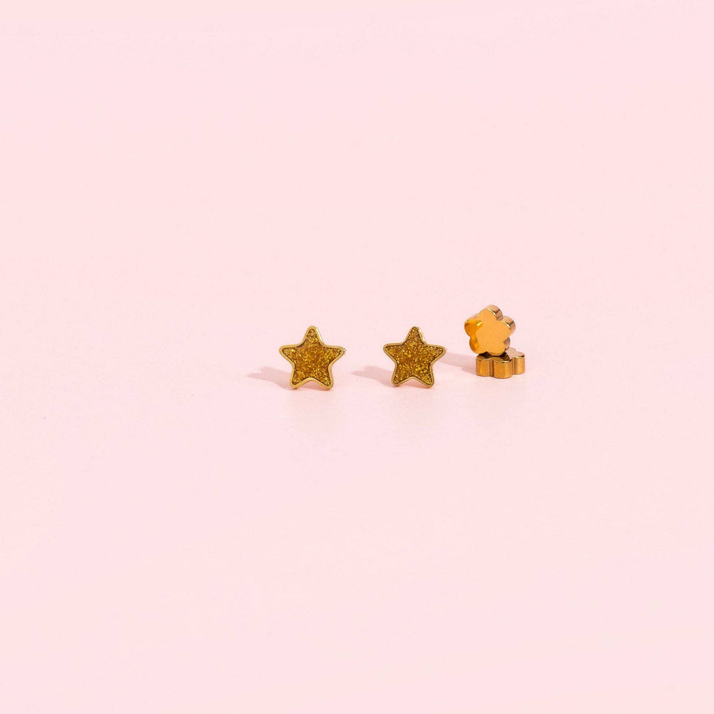 A pair of small gold star stud earrings, with one shown from the front and the other from the side, on a solid pale pink background.