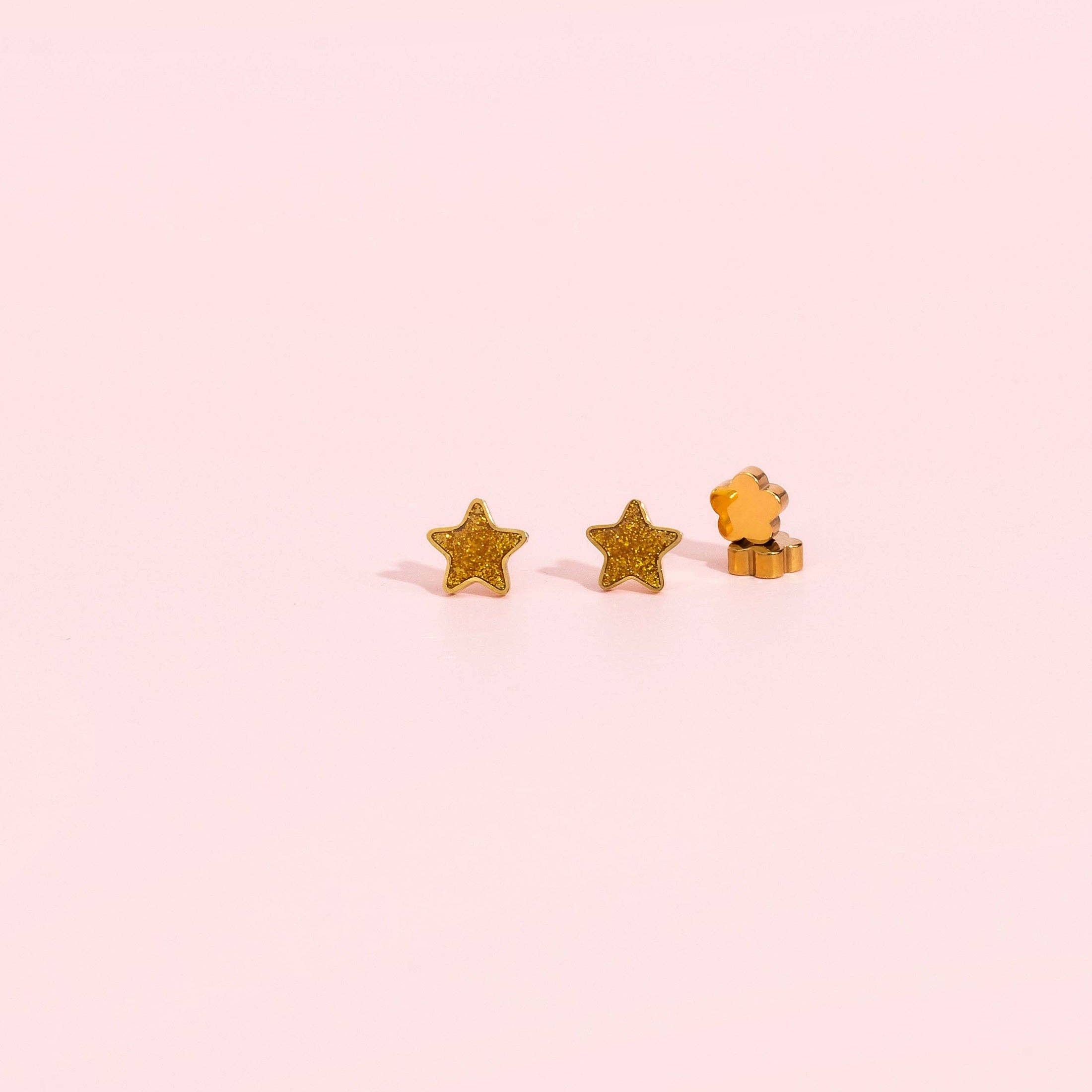 A pair of small gold star stud earrings, with one shown from the front and the other from the side, on a solid pale pink background.
