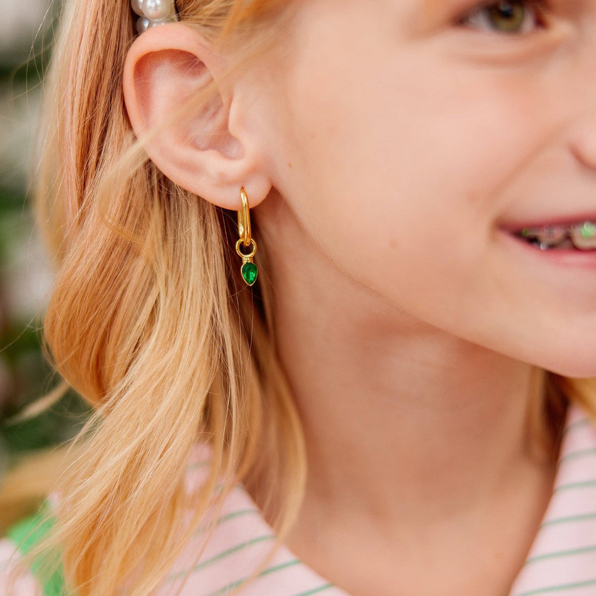 A close-up lifestyle shot of a young girl wearing a delicate gold huggie hoop earring with a small green teardrop gemstone dangle.