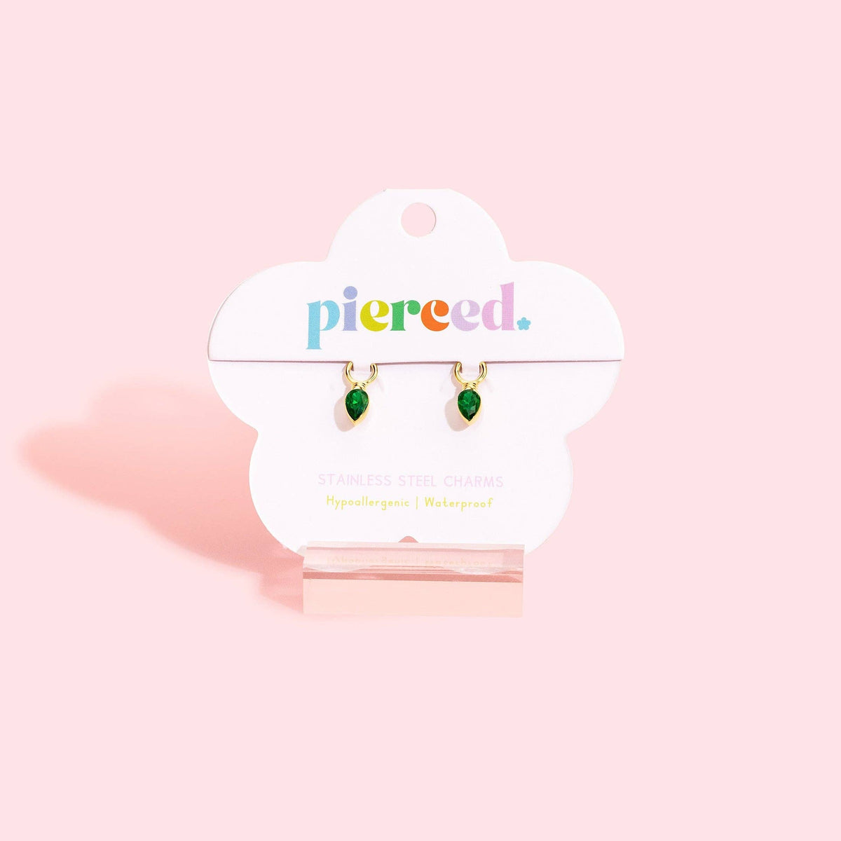 A pair of small teardrop-shaped green charm earrings on a white cloud-shaped display card with the brand name 'piered.' in colorful lowercase letters, set against a solid light pink background.