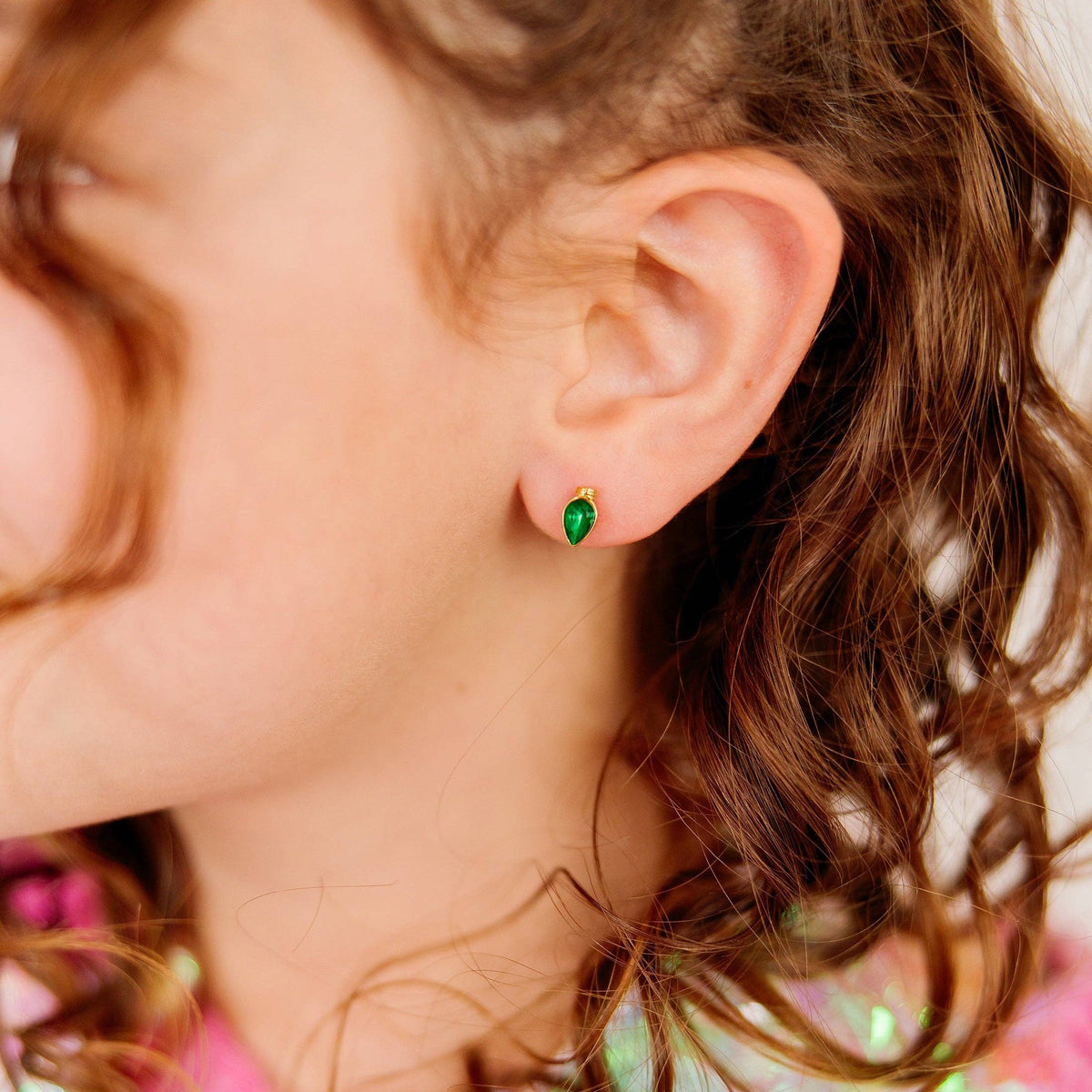 Close-up of a child's ear wearing a small gold stud earring with a green marquise-cut gemstone.