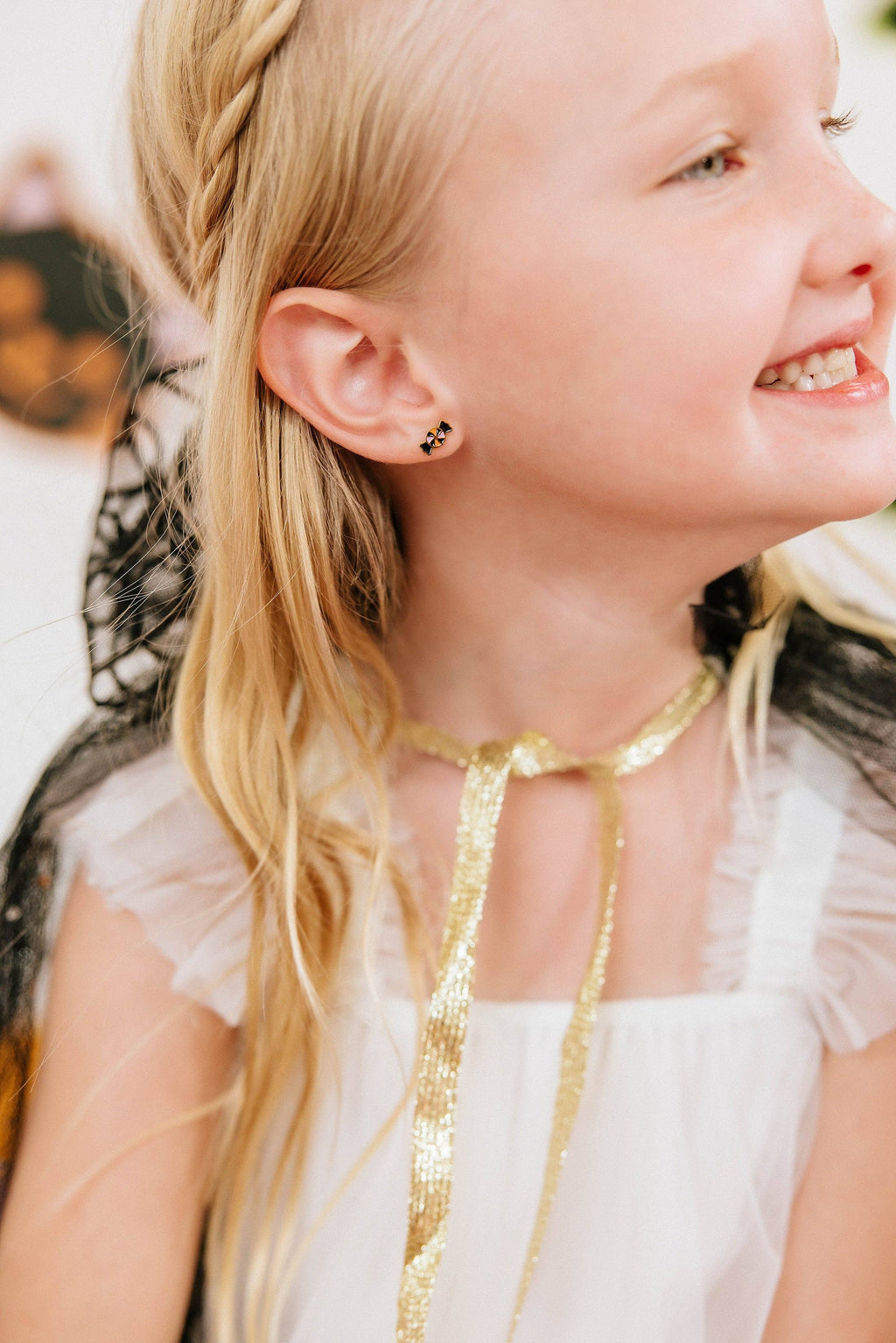 A close-up lifestyle shot of a young girl with blonde hair wearing a white dress and a gold ribbon choker, featuring a small black and gold spider stud earring on her ear.
