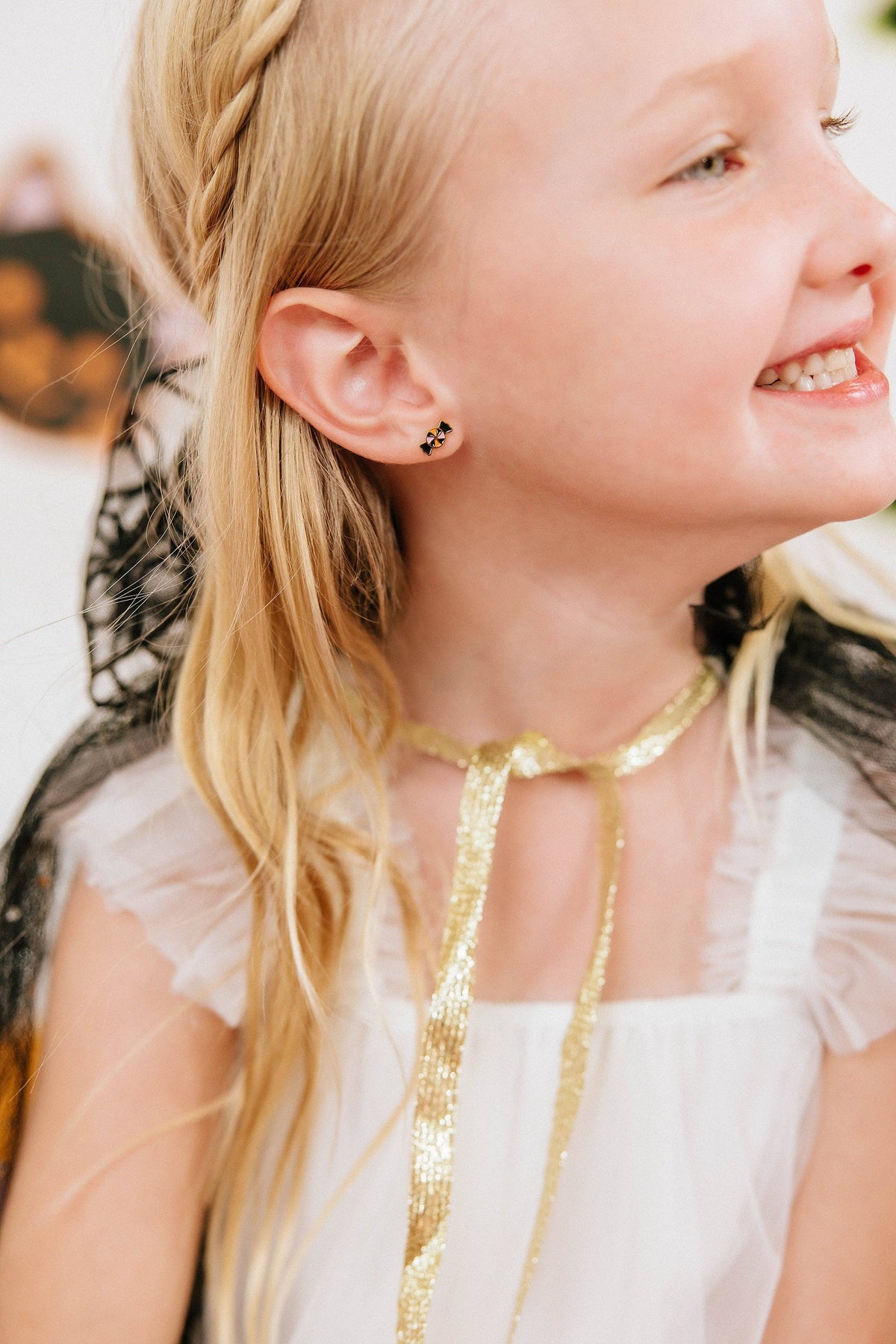 A close-up lifestyle shot of a young girl with blonde hair wearing a white dress and a gold ribbon choker, featuring a small black and gold spider stud earring on her ear.