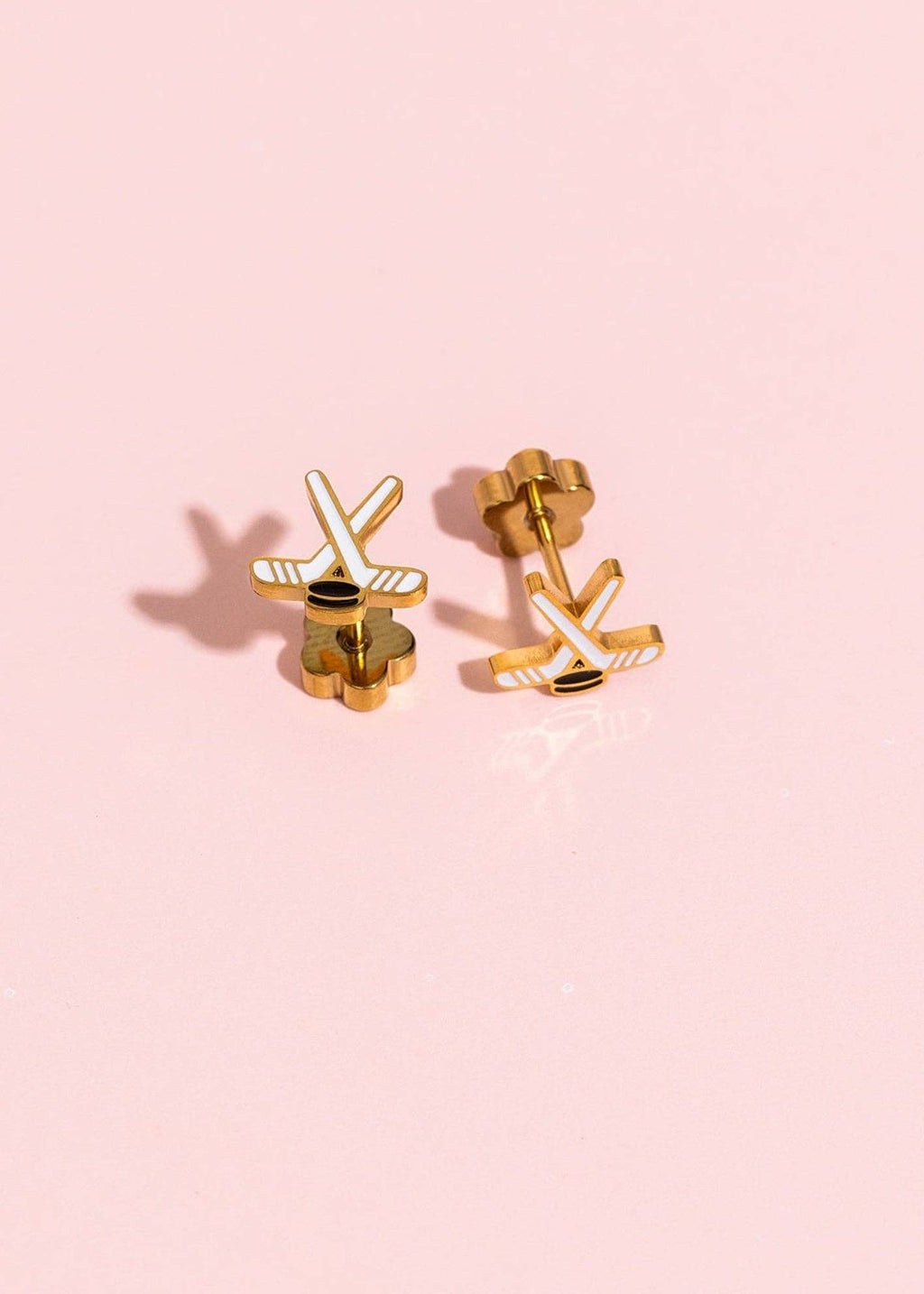 A pair of gold-toned stud earrings shaped like crossed hockey sticks with white enamel details, displayed on a soft pink background.