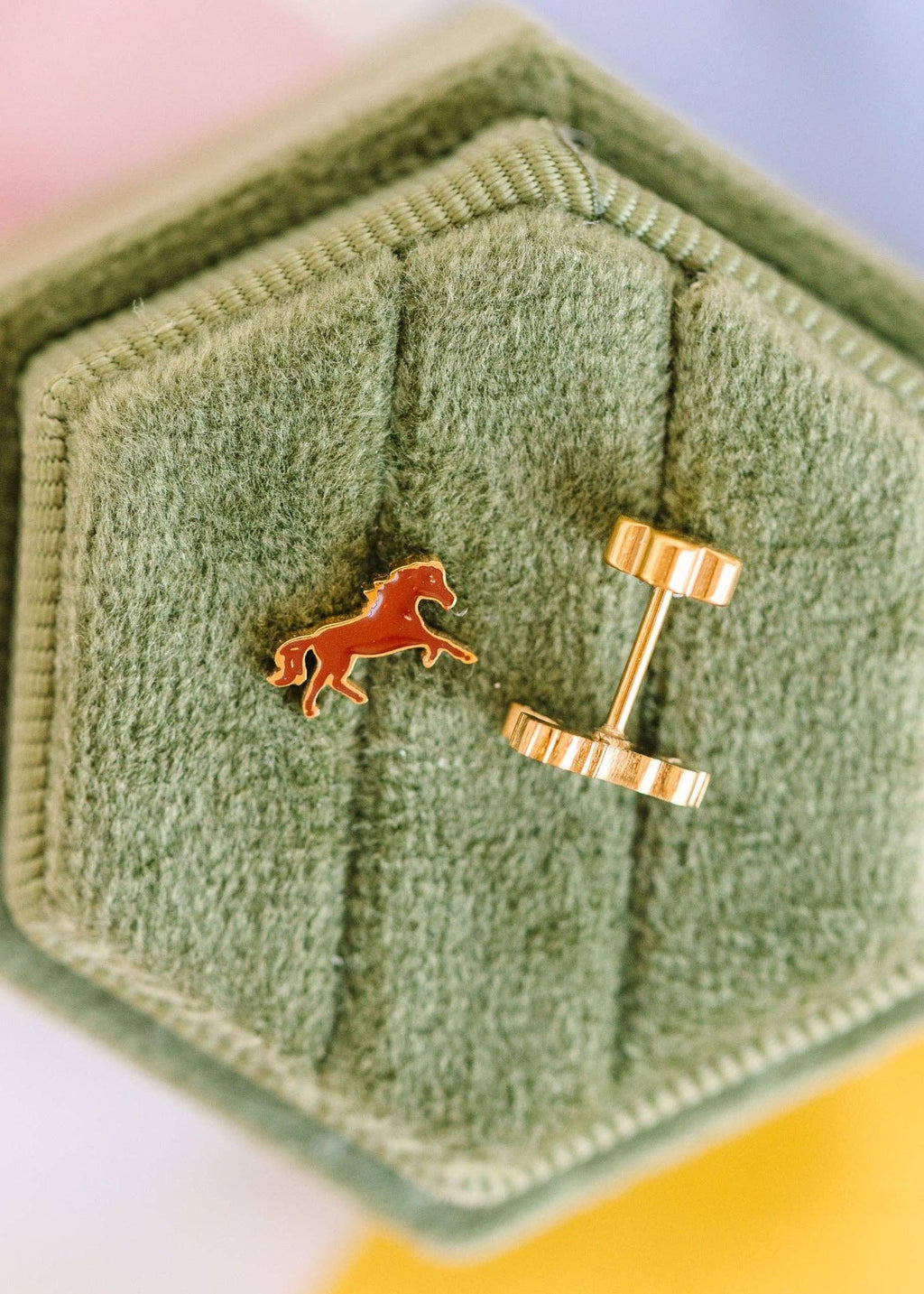 A close-up of a pair of gold cufflinks displayed on a textured sage green hexagonal jewelry box. One cufflink is shown from the front, featuring a small orange horse emblem, and the other is turned to show the gold post and loop.