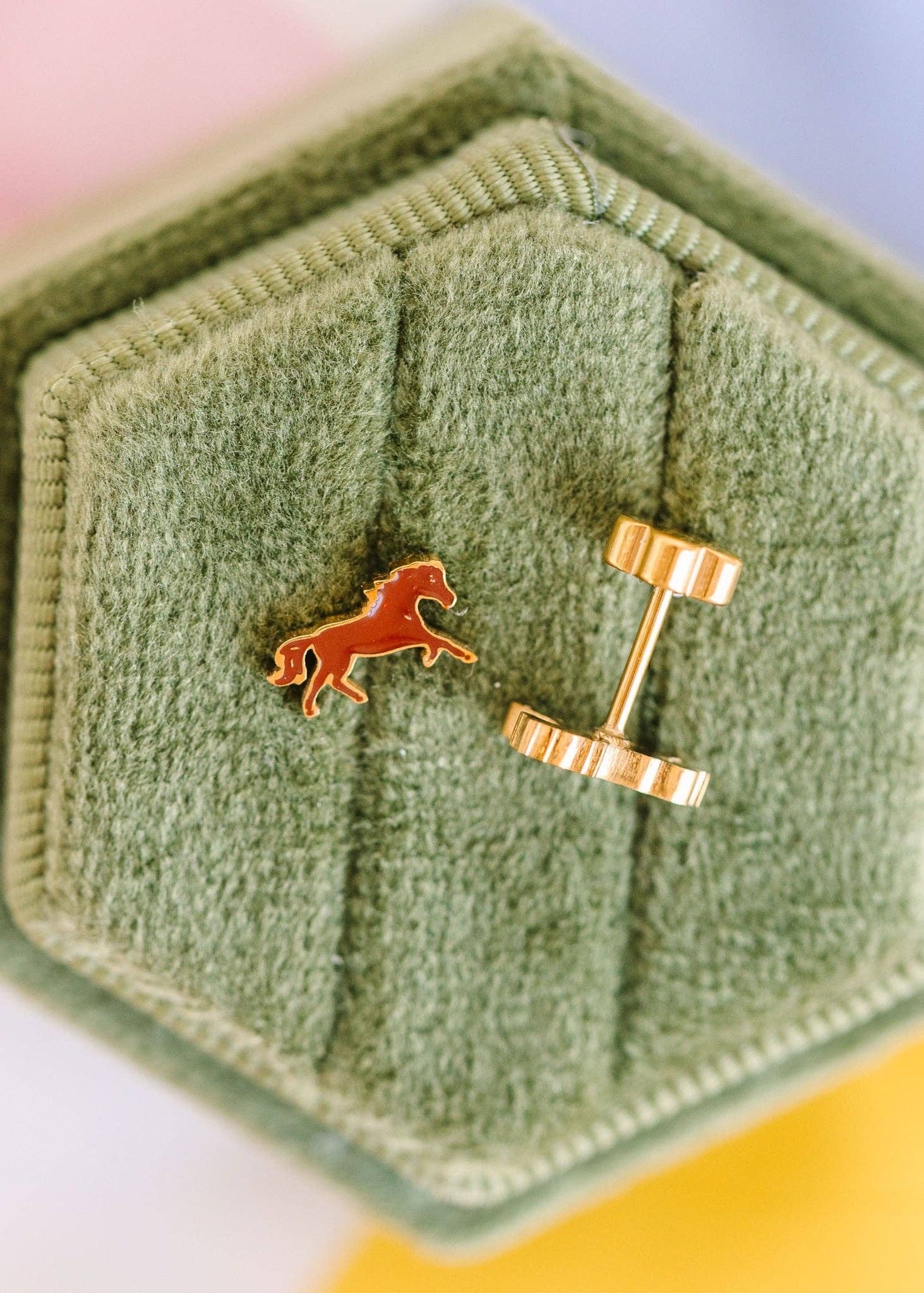 A close-up of a pair of gold cufflinks displayed on a textured sage green hexagonal jewelry box. One cufflink is shown from the front, featuring a small orange horse emblem, and the other is turned to show the gold post and loop.