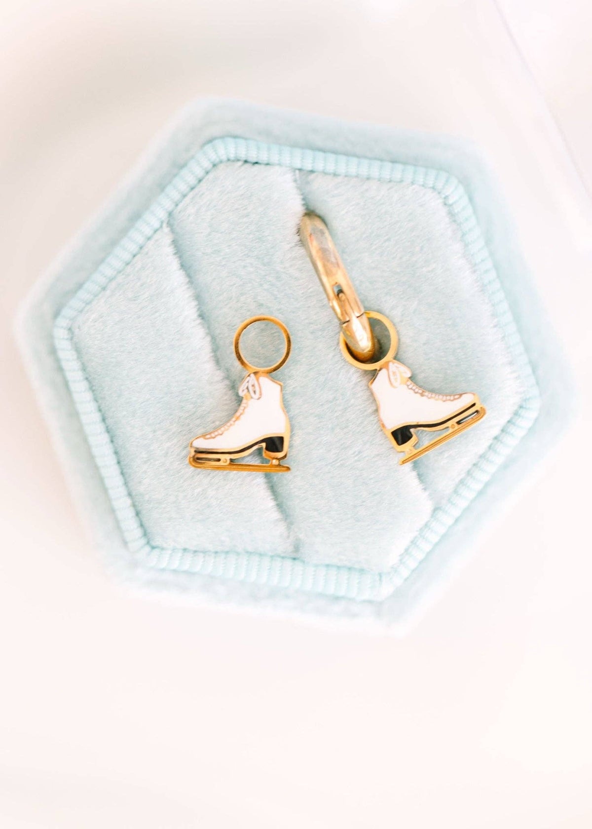 A pair of gold-tone ice skate charms with white enamel detailing, displayed on a light blue hexagonal jewelry box insert against a white background.