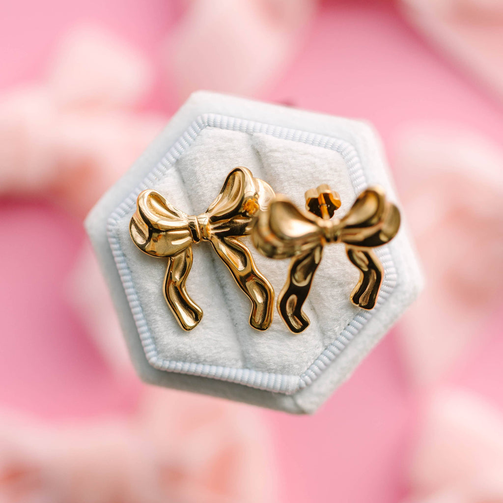 Gold bow-shaped earrings on a white hexagonal stand with a pink floral background