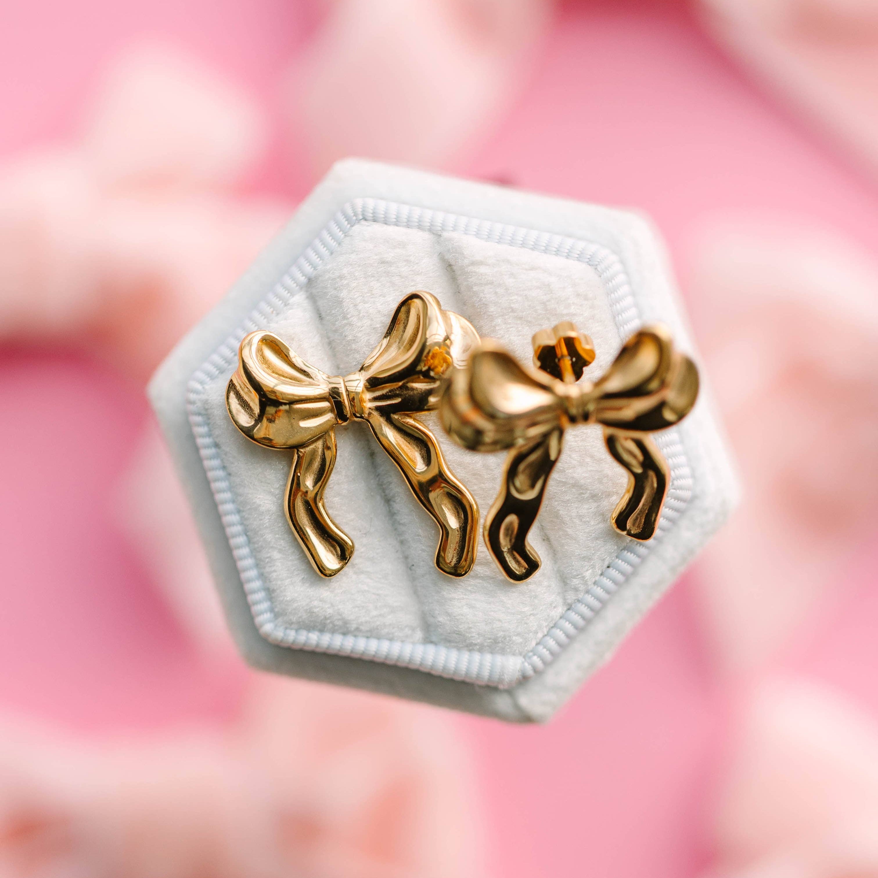 Gold bow-shaped earrings on a white hexagonal stand with a pink floral background