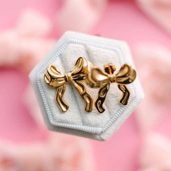 Gold bow-shaped earrings on a white hexagonal stand with a pink floral background
