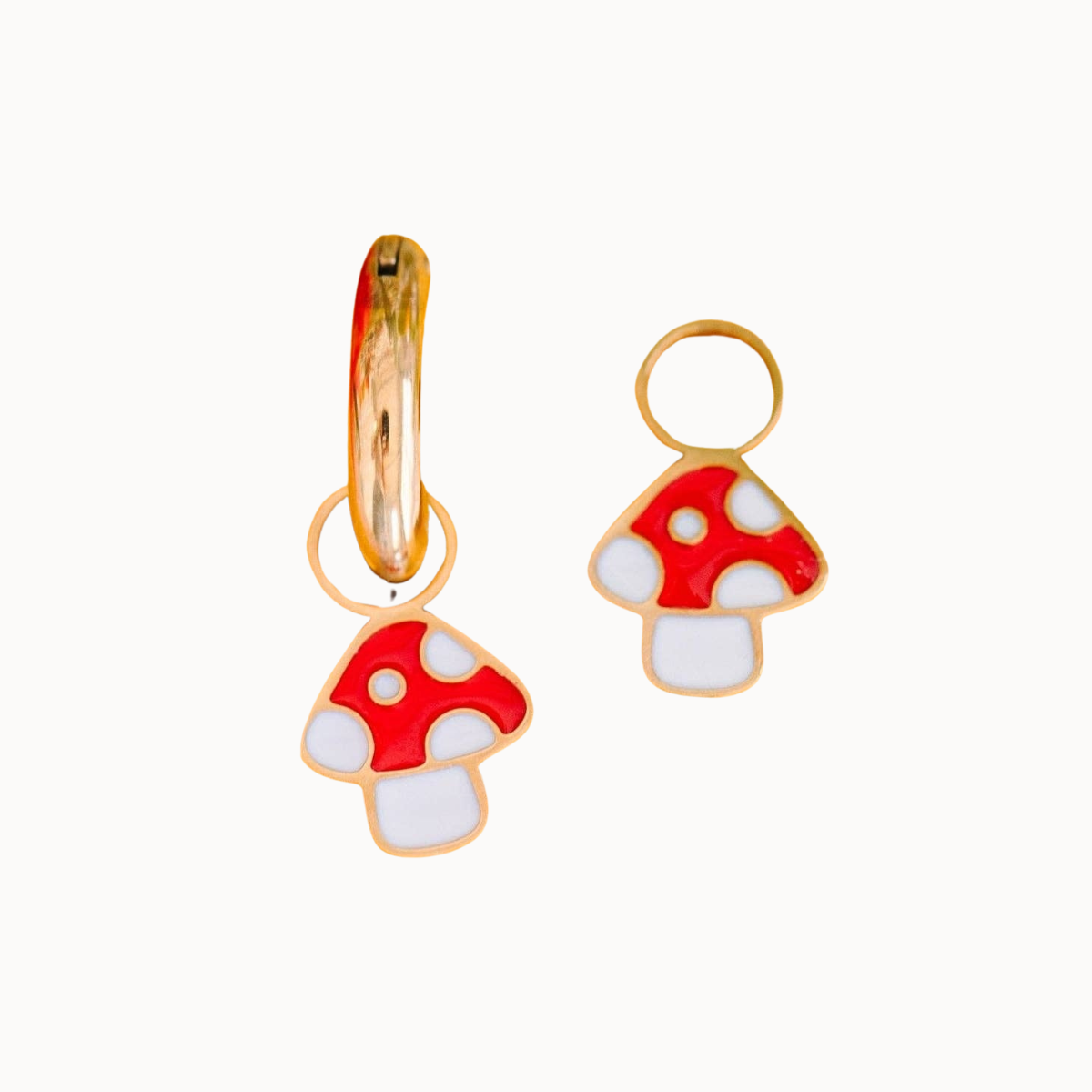 A pair of gold hoop earrings with dangling mushroom charms. The mushroom charms feature a red cap with white spots and a white stem, outlined in gold.