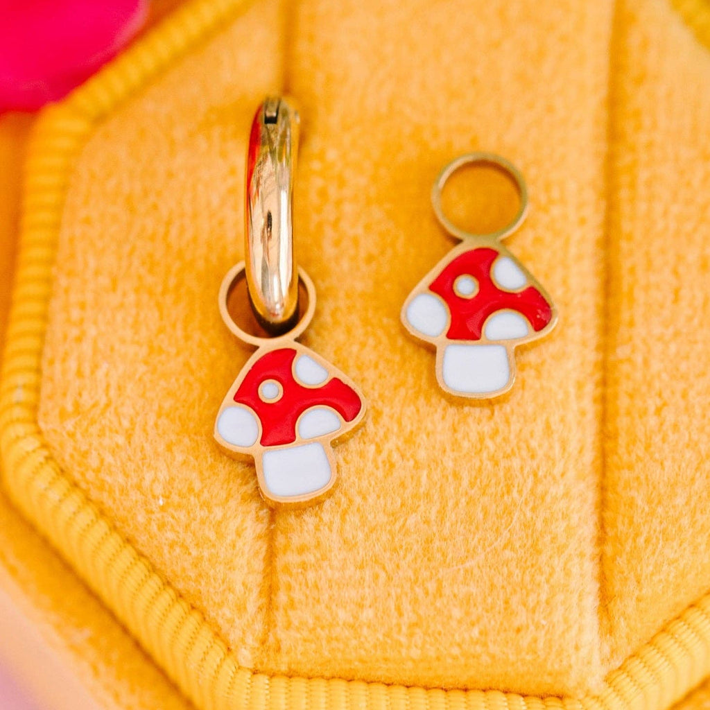 A pair of small mushroom-shaped earrings with red and white enamel details and gold-tone metal rings, displayed on a yellow textured jewelry stand.