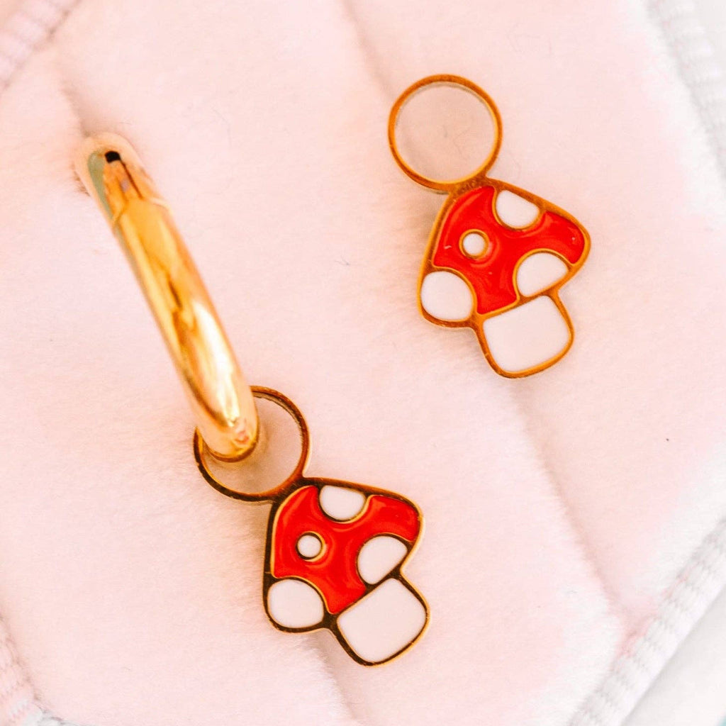 A pair of gold hoop earrings with red and white mushroom charms, displayed on a light pink hexagonal jewelry box.