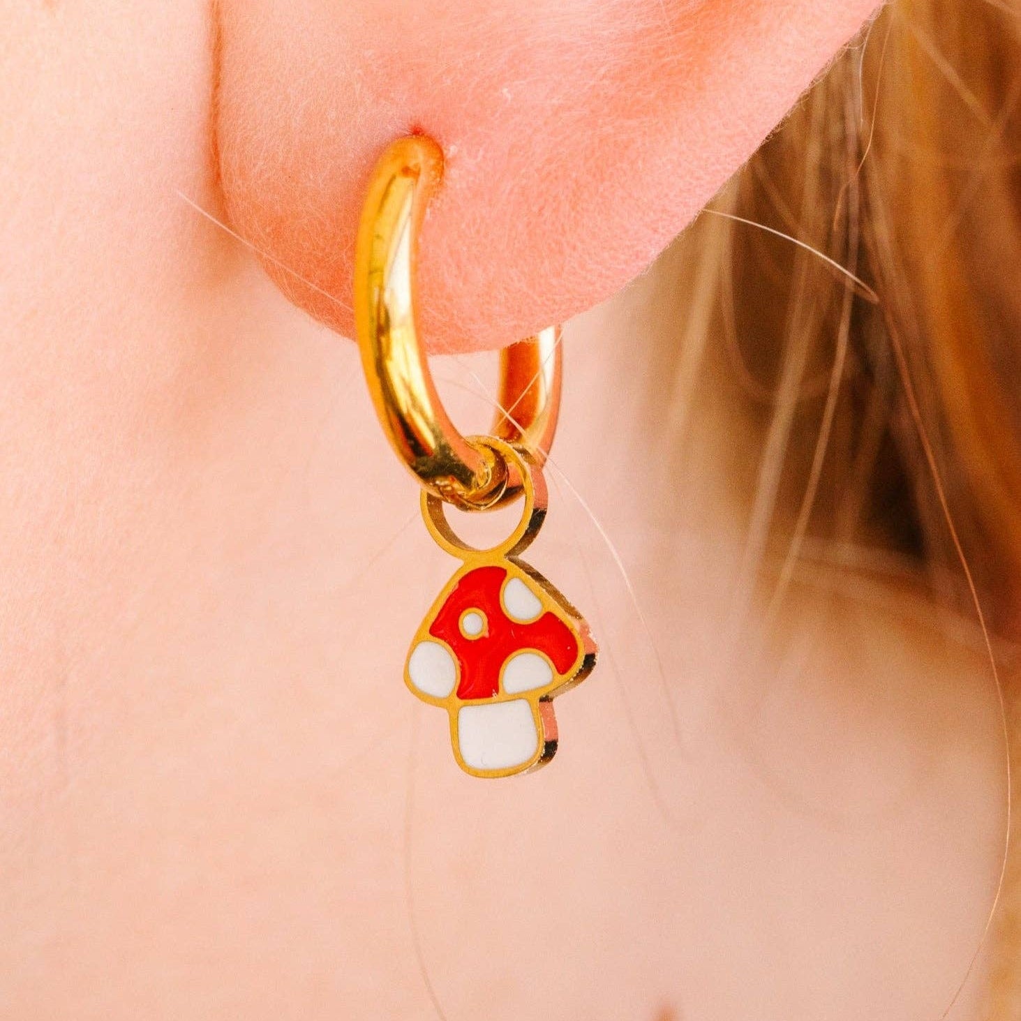 A close-up of a gold hoop earring worn on a person's ear, featuring a dangling mushroom charm with red and white enamel.