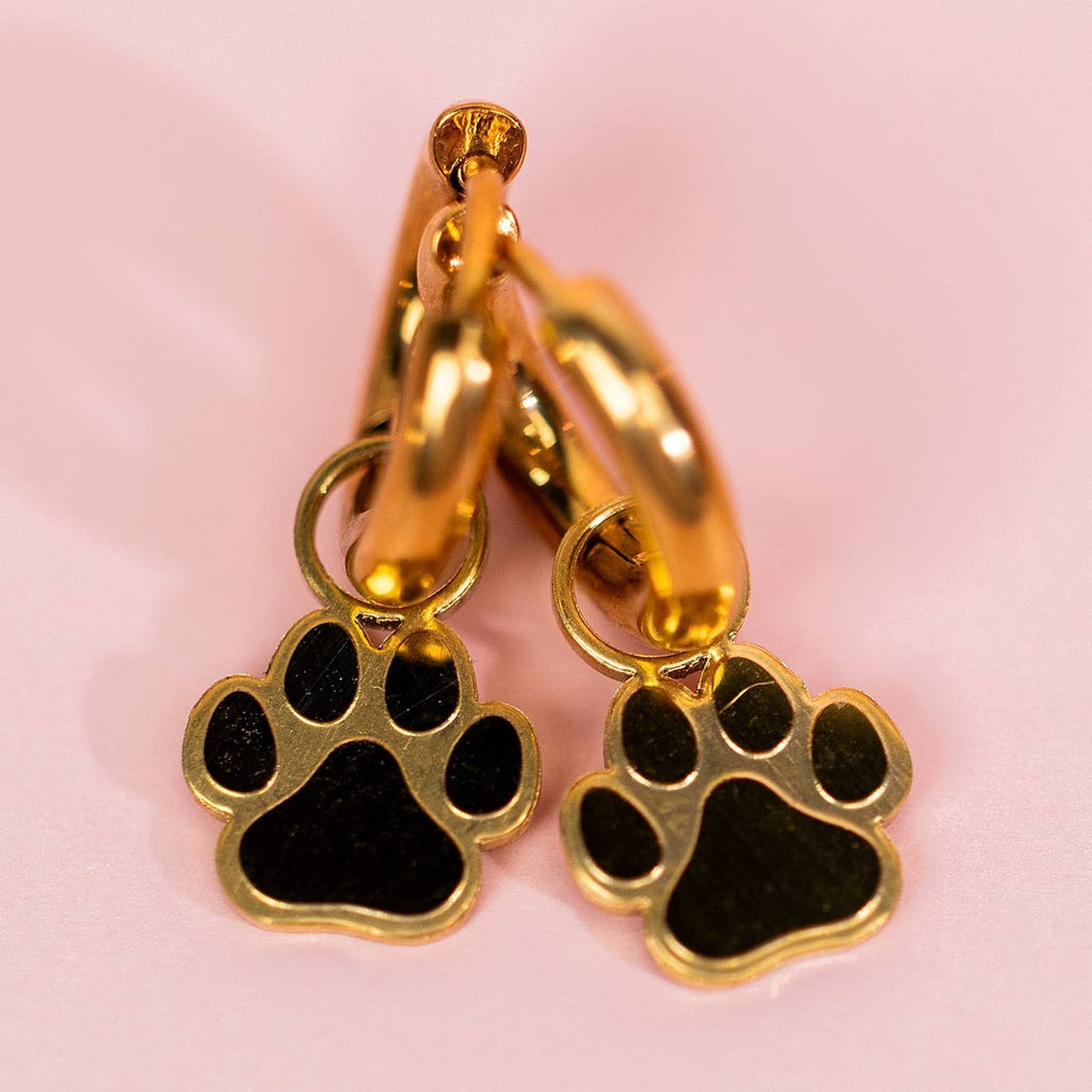Gold hoop earrings with black paw print charms on a pink background