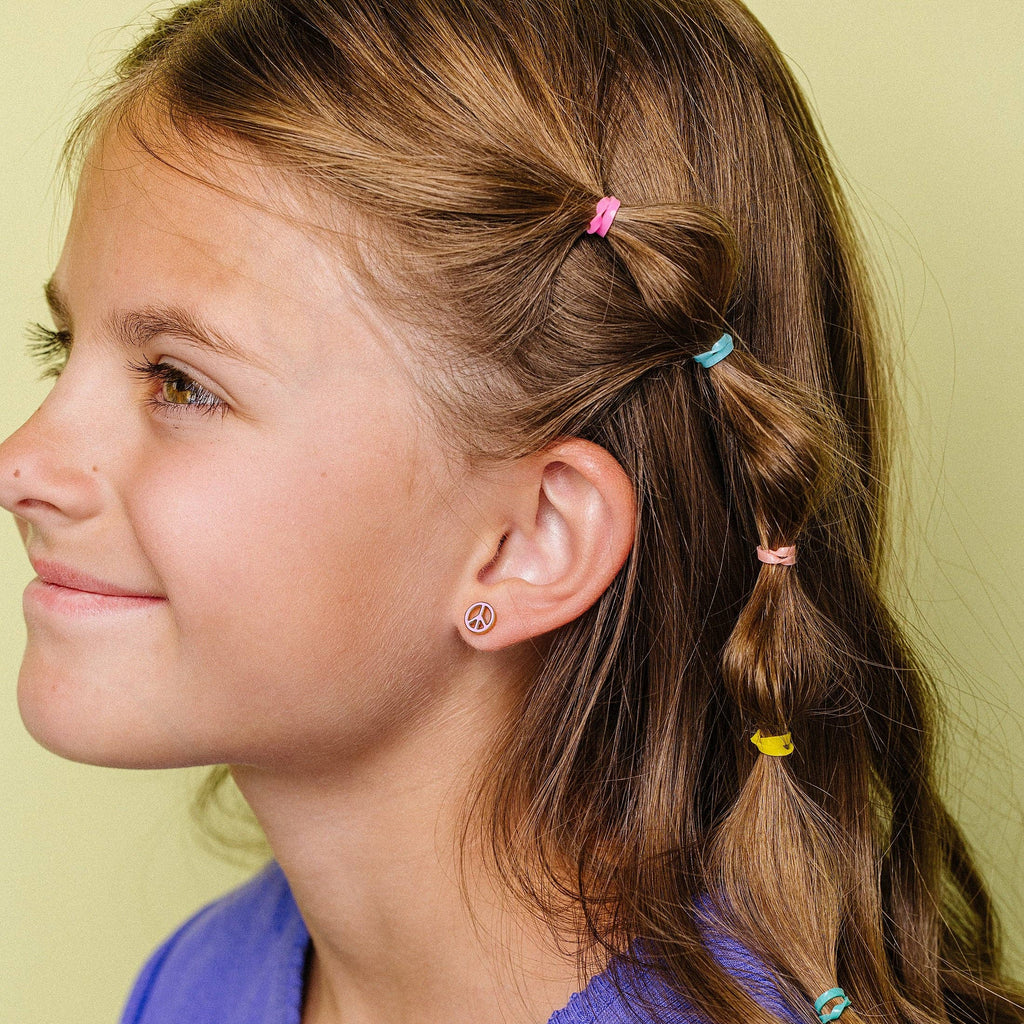 A young girl with light brown hair styled in a side braid with colorful elastic hair ties, wearing a purple ribbed shirt against a pale yellow-green background.