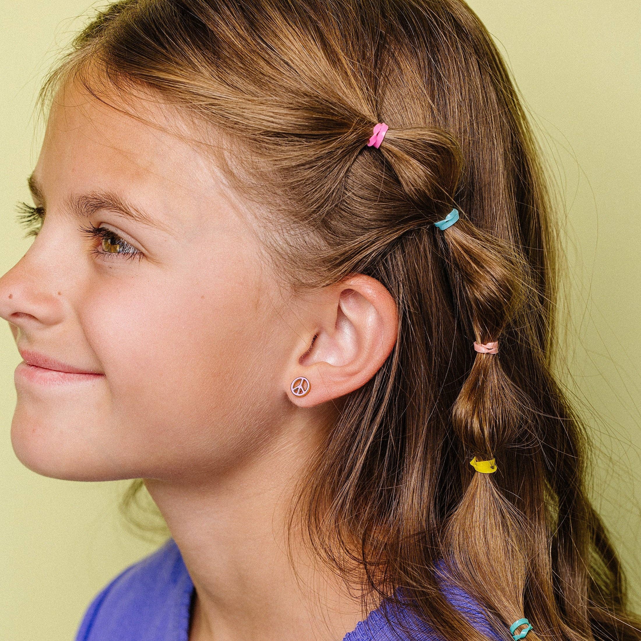 A young girl with light brown hair styled in a side braid with colorful elastic hair ties, wearing a purple ribbed shirt against a pale yellow-green background.