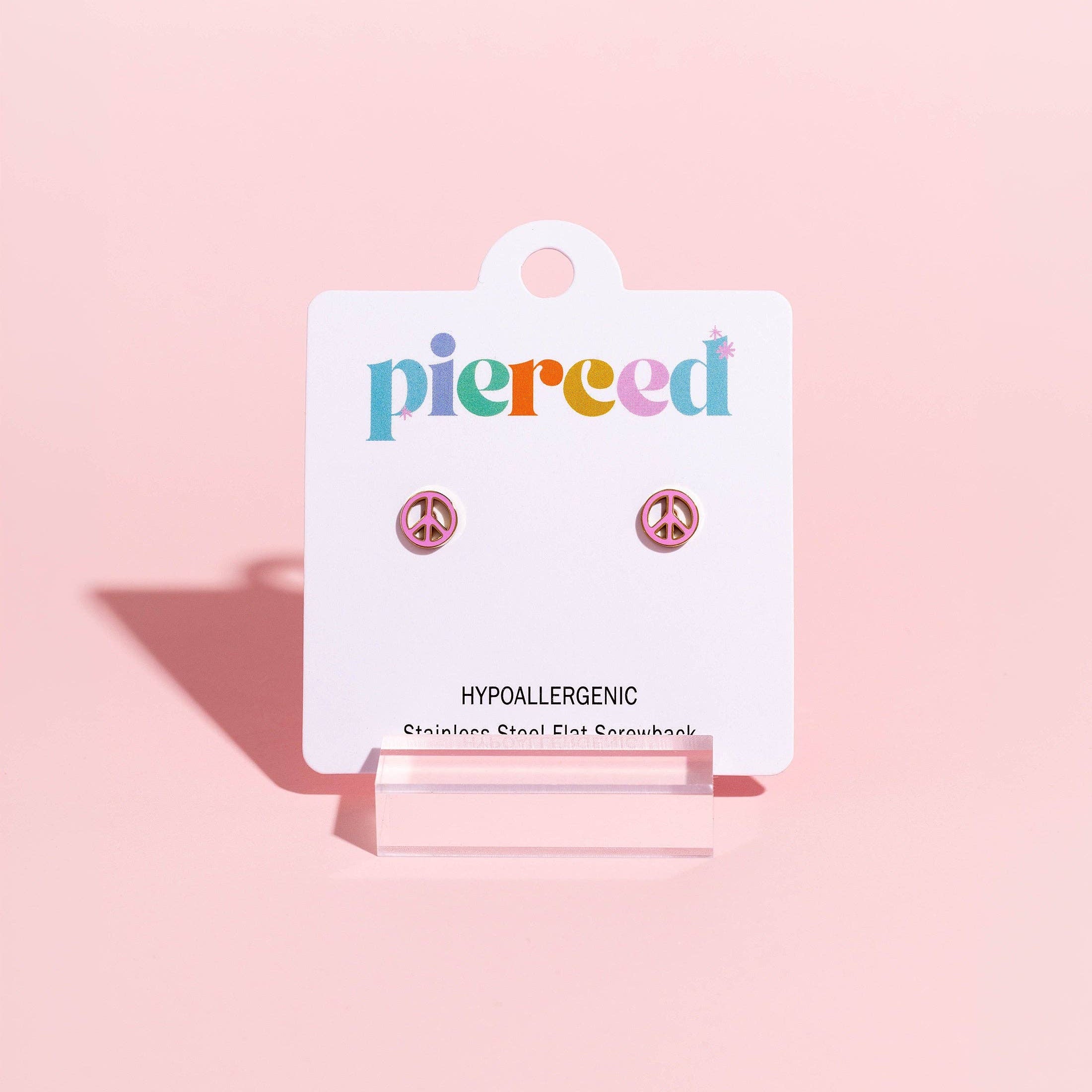 A pair of small stud earrings displayed on a white Pierced brand card, set against a solid pastel pink background.