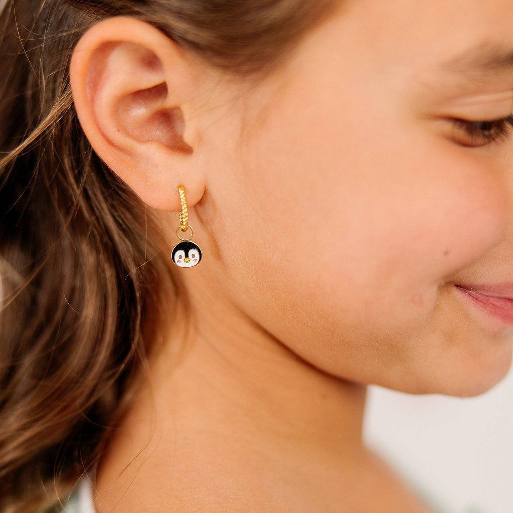 A close-up side profile of a young girl wearing a gold huggie hoop earring with a dangling penguin charm.