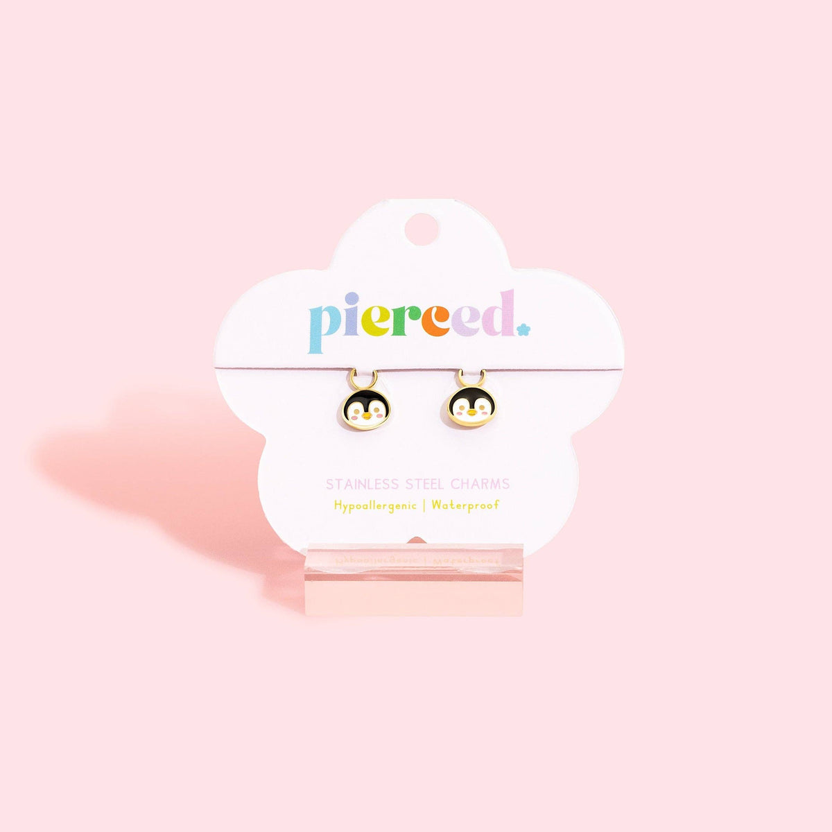 A pair of small penguin-shaped stud earrings displayed on a white cloud-shaped Pierceed brand card, set against a solid pastel pink background.