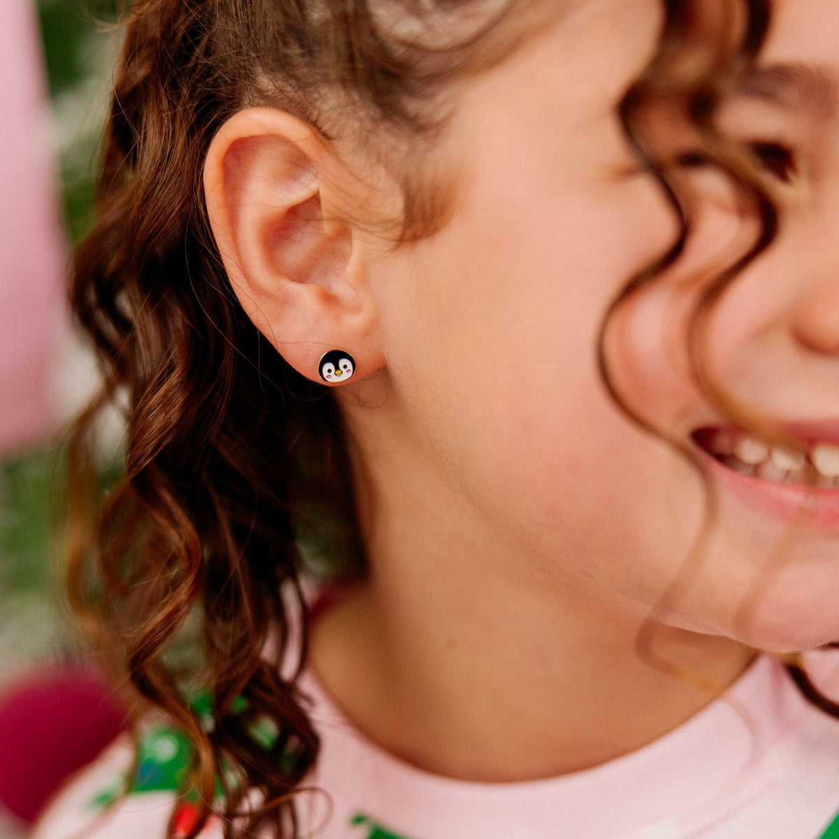 A close-up of a young girl wearing a small black and white penguin stud earring, with her curly brown hair and smiling face visible.