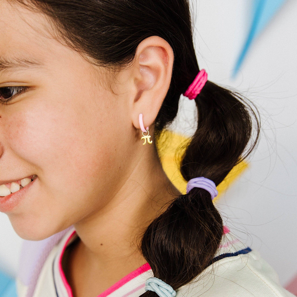 A close-up side profile of a smiling girl with dark brown hair in a ponytail, wearing a small gold Greek letter 'pi' earring. She is wearing a white V-neck top with pink and navy trim, and her hair is secured with colorful pastel hair ties.