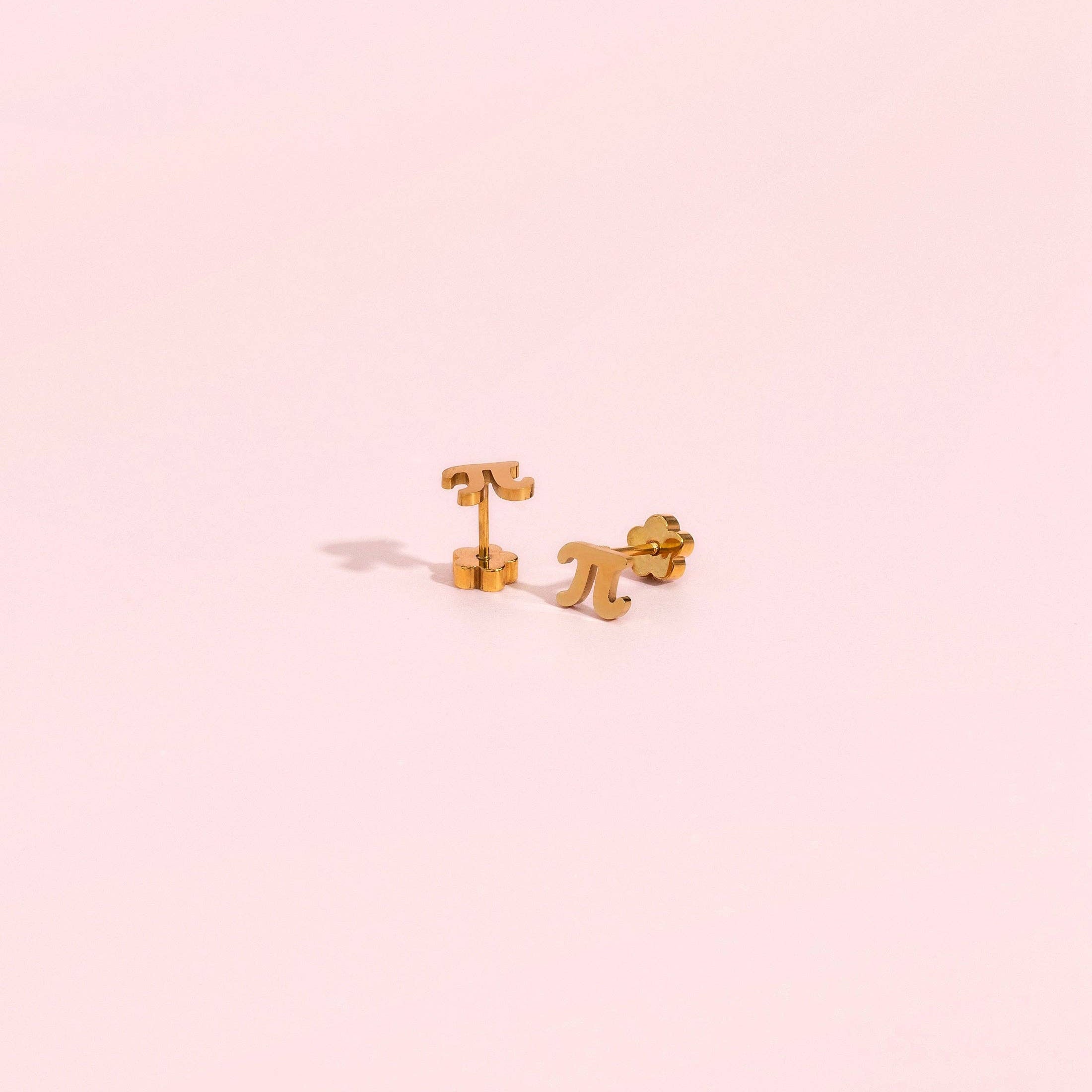 A pair of small gold stud earrings shaped like a stylized letter T and a stylized letter P, displayed on a solid pale pink background.