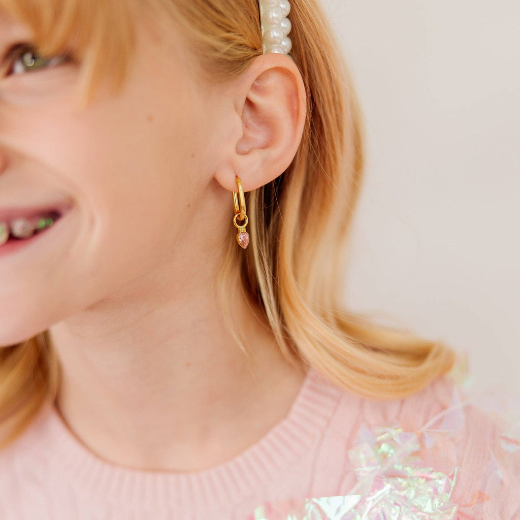A close-up of a young girl wearing a delicate gold huggie hoop earring with a small pink bead dangle, paired with a pearl hair clip and a pink top.