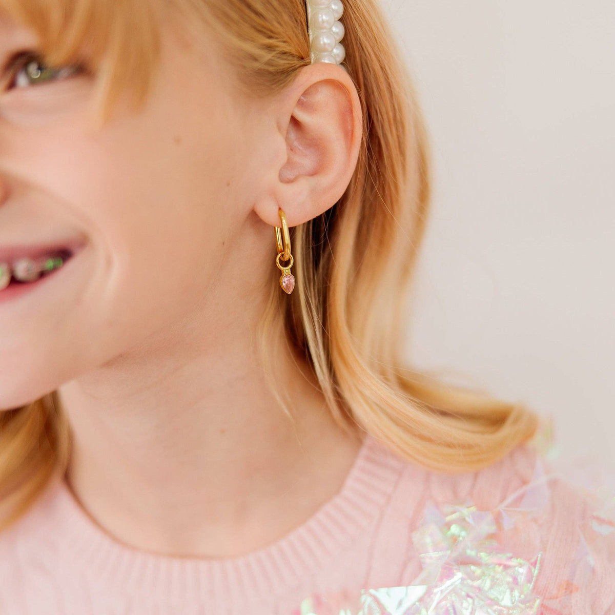A close-up of a young girl wearing a delicate gold huggie hoop earring with a small pink bead dangle, paired with a pearl hair clip and a pink top.