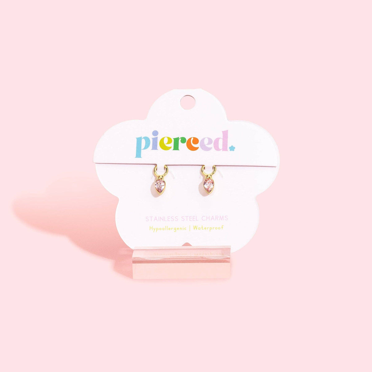 A pair of gold-toned steel charm earrings with clear teardrop-shaped charms, displayed on a white cloud-shaped card labeled 'pierceed.' against a soft pink background.