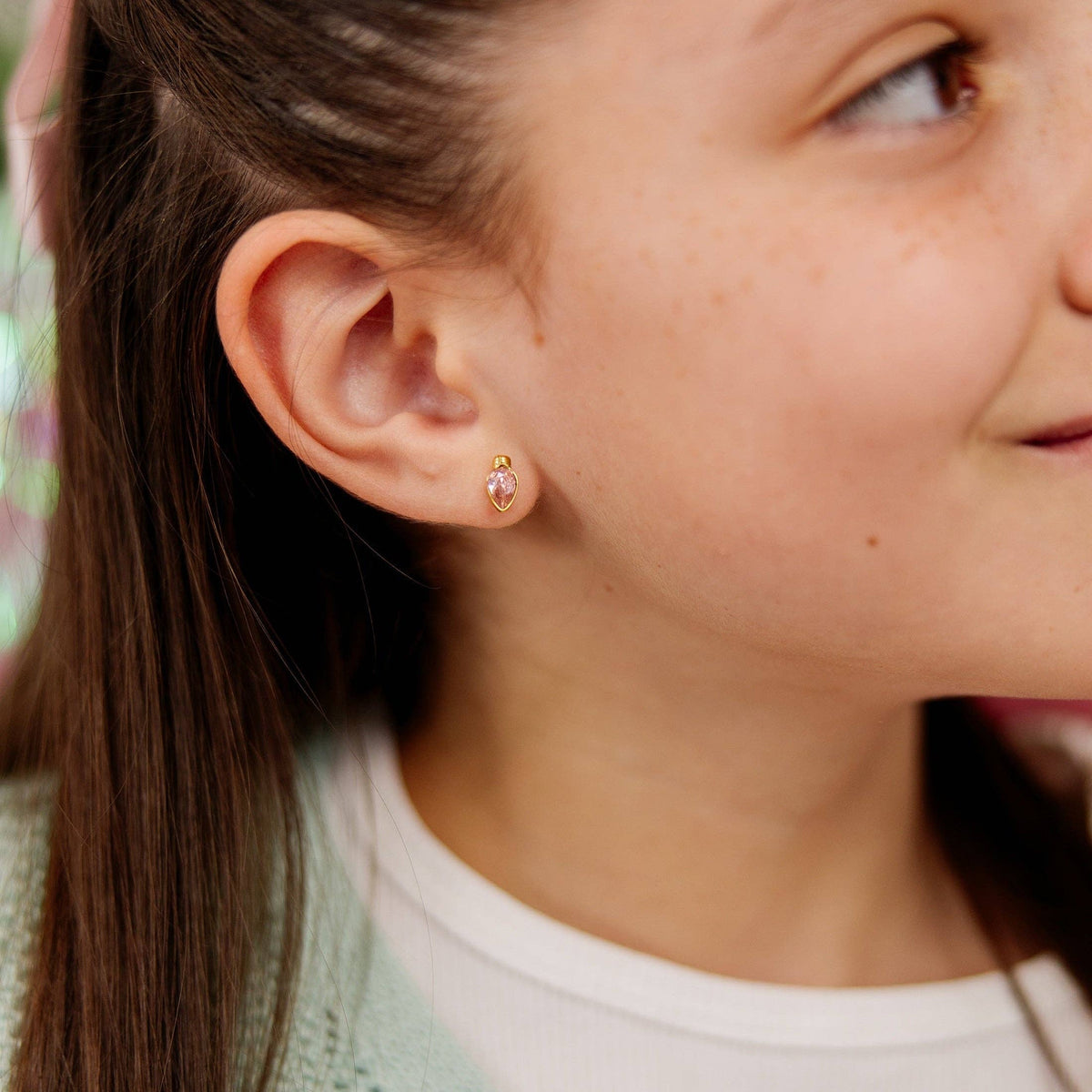 A close-up of a young girl's ear wearing a small gold stud earring with a pink christmas light-shaped crystal, set against her face and hair.