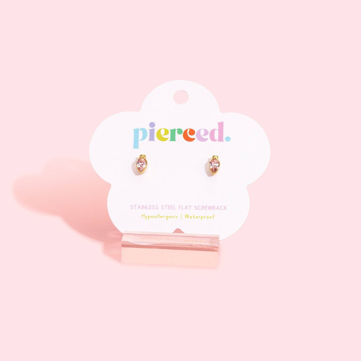 A pair of small flat screwback earrings displayed on a white cloud-shaped card stand against a soft pink background. The card shows the brand name 'pierce.